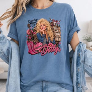 May include: A blue t-shirt featuring a graphic print of a blonde woman in a denim jacket, a guitar, pink cowboy boots, a pink hat, and a cow skull. The text includes Country Roads, Jolene, 9 to 5, I Will Always Love You, Coat of Many Colors, and Dolly.