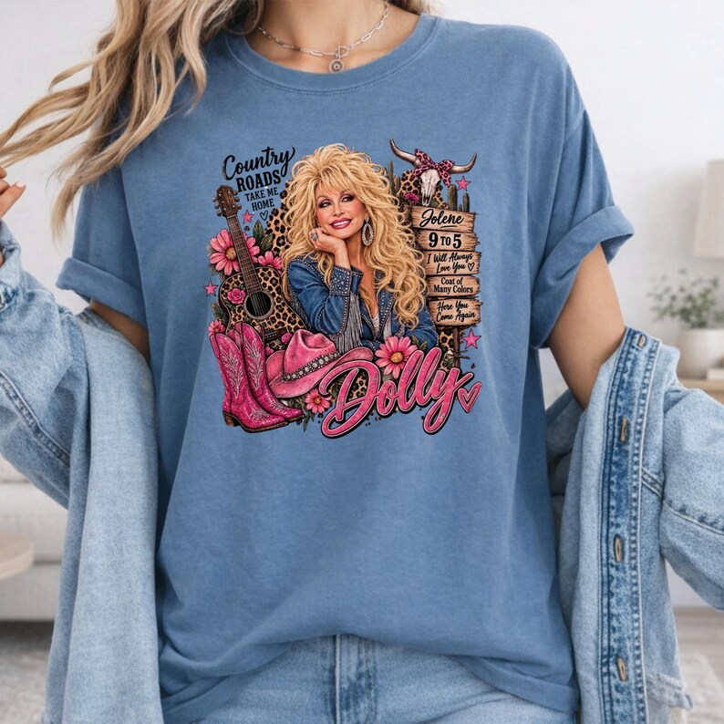 May include: A blue t-shirt featuring a graphic print of a blonde woman in a denim jacket, a guitar, pink cowboy boots, a pink hat, and a cow skull. The text includes Country Roads, Jolene, 9 to 5, I Will Always Love You, Coat of Many Colors, and Dolly.