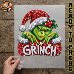 May include: A DTF transfer sheet featuring a green character wearing a red and white Santa hat adorned with red berries and green leaves. The character has a mischievous expression and is positioned above the white text GRINCH on a red background.