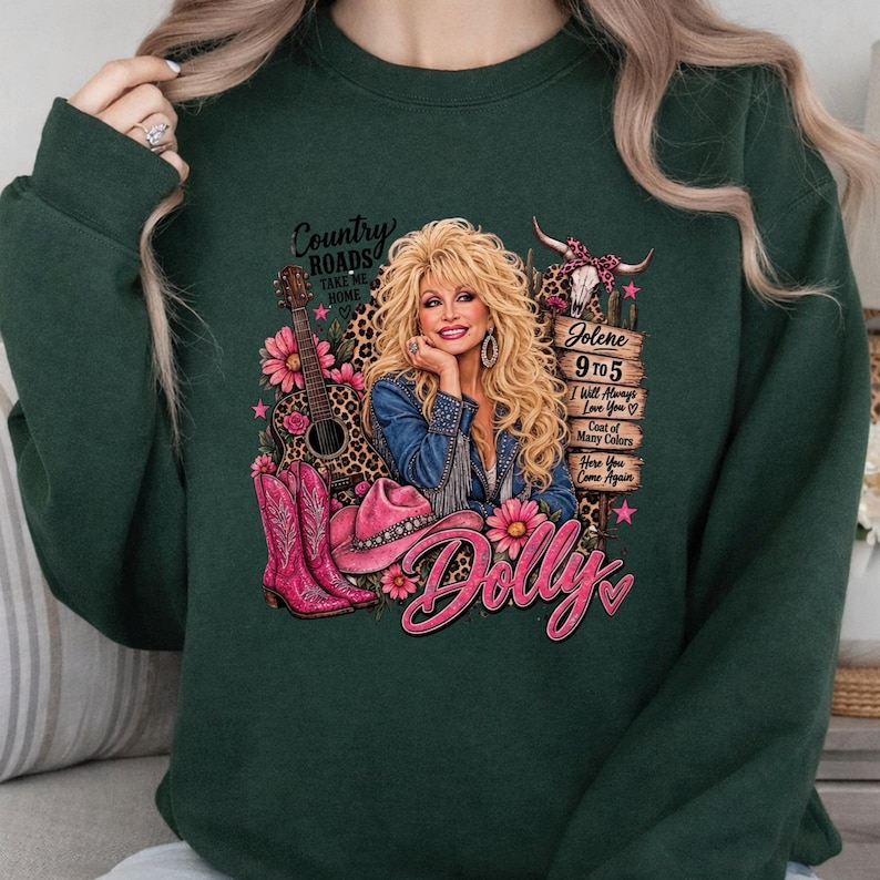 May include: A dark green sweatshirt featuring a graphic print of a blonde woman in a denim jacket, a guitar, pink cowboy boots, a pink hat, and a cow skull. The text includes Country Roads, Jolene, 9 to 5, I Will Always Love You, and Dolly.