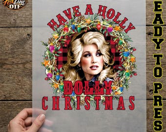 May include: A DTF transfer sheet featuring a portrait of Dolly Parton surrounded by a Christmas wreath with red, green, and blue lights, pinecones, and stars. The text reads HAVE A HOLLY DOLLY CHRISTMAS in red, with a red and black plaid background.