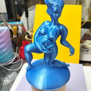 Può includere: Una statuetta stampata in 3D di colore blu metallizzato raffigurante una figura inginocchiata con capelli fluenti, che tiene in mano un piccolo oggetto sferico. La figura poggia su una base circolare blu sopra un supporto cilindrico in legno.