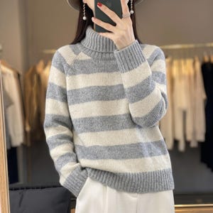 May include: A long-sleeved sweater featuring a horizontal striped pattern in alternating shades of light gray and off-white. The garment is designed with a ribbed turtleneck collar, ribbed cuffs at the wrists, and a ribbed hemline at the waist.