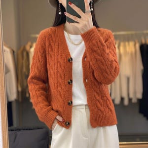May include: A burnt orange cable-knit cardigan featuring a V-neckline, long sleeves, and four dark brown buttons down the front. The knit garment is worn over a plain white t-shirt and is paired with white trousers, showcasing a textured pattern.