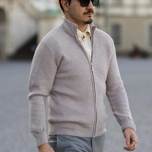 Può includere: Un maglione con zip in maglia a coste color beige chiaro, caratterizzato da un collo alto, maniche lunghe con polsini a coste e una cerniera frontale a tutta lunghezza. Il maglione è indossato sopra una camicia a colletto chiaro, abbinato a pantaloni grigi e occhiali da sole scuri.