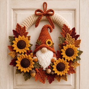 Può includere: Una ghirlanda all'uncinetto con una base circolare beige e un fiocco superiore, decorata con tre girasoli gialli, varie foglie dai colori autunnali arancioni, rossi e verdi, piccole ghiande e un gnomo centrale con cappello arancione e barba bianca.