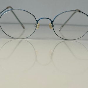 Op de afbeelding: Een blauwe metalen bril met ovaalvormige glazen en dunne draadframes. De bril is voorzien van heldere glazen, verstelbare neuspads en slanke pootjes die vanaf de zijkanten van het frame naar buiten steken voor een lichtgewicht uitstraling.