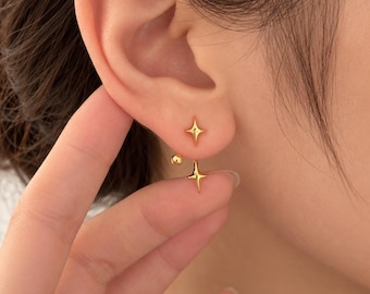 May include: A gold-toned earring featuring two four-pointed star shapes. The smaller star sits on the front of the earlobe, while the larger star hangs from the back, connected by a thin metal post with a small spherical backing.
