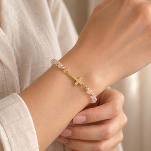 May include: A beaded bracelet featuring small, pale pink round beads interspersed with tiny gold-toned metallic spheres. The bracelet includes a central gold-toned cross charm, positioned horizontally along the strand of beads on the wrist.