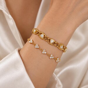 May include: A set of two gold-toned bracelets. The top bracelet is a thick chain link style featuring embedded triangular clear stones. The bottom bracelet is a delicate chain featuring small clusters of three clear stones spaced evenly apart.