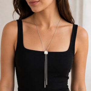 May include: A silver-toned lariat necklace featuring a central, smooth, white oval-shaped stone set in a metallic frame. Two thin silver chains extend downward from the stone, each ending in a slender, cylindrical silver-toned metal bar pendant.