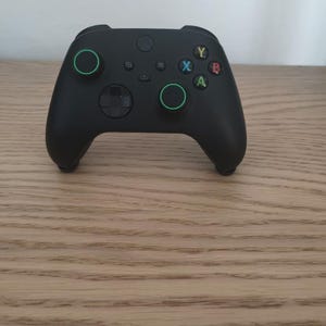 May include: A matte black wireless video game controller featuring two analogue sticks with green circular accents, a directional pad, and four primary action buttons labelled Y, X, B, and A in yellow, blue, red, and green colours respectively.