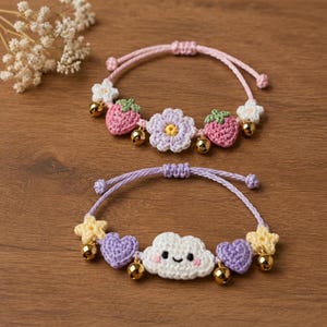 Pode incluir: Duas pulseiras de cordão ajustável com pingentes de crochê. A superior tem morangos rosa, uma flor lavanda, flores brancas e guizos dourados. A inferior apresenta uma nuvem branca sorridente, corações roxos, estrelas amarelas e guizos dourados.