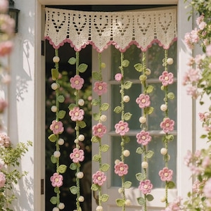 May include: A crochet door curtain featuring a cream-coloured valance with a heart pattern and scalloped edges. Multiple vertical strands hang from the valance, decorated with green crocheted leaves, small white spheres, and pink five-petaled flowers.