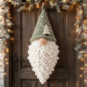Puede incluir: Un adorno de pared de gnomo tejido a ganchillo con un gorro puntiagudo verde salvia, patrones de cruces blancas y una aplicación de árbol color crema. El gnomo tiene una nariz redonda color melocotón y una larga barba blanca de pompones.