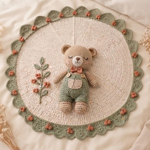 May include: A round, cream-colored crocheted mat featuring a scalloped sage green border adorned with small peach-colored clusters. A crocheted teddy bear wearing sage green overalls and a peach bow tie sits on the mat next to a crocheted floral sprig.