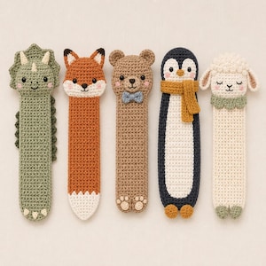May include: Five crochet bookmarks shaped like animals: a green triceratops, an orange fox, a brown bear with a blue bow tie, a black and white penguin with a yellow scarf, and a cream-colored sheep with a green collar, all featuring embroidered faces.