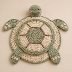 May include: A crocheted turtle mat featuring a sage green and cream colour palette. The turtle has a round shell with geometric segments, four flippers with cream-coloured accents, and a head with embroidered closed eyes and pink cheeks.