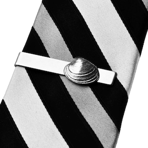 May include: A silver tie clip featuring a detailed seashell design. The clip is rectangular and holds a black, white, and gray striped tie. The seashell is the focal point, adding a nautical touch to the accessory.