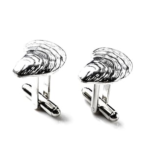 以下が含まれることがあります： 牡蠣の貝殻の形をした銀製のカフスボタンのペア。カフスボタンは、詳細で質感のあるデザインになっています。