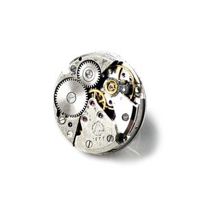 May include: Close-up of a silver watch movement with visible gears and a small, round, gold-colored part. The movement is exposed and shows the intricate workings of the watch.