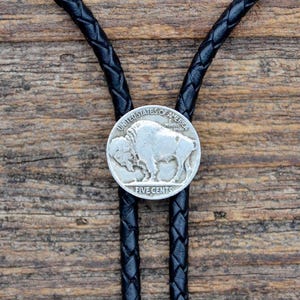 Könnte beinhalten: Ein schwarzes geflochtenes Leder-Boloneckband mit einer silbernen Büffel-Nickel-Münze als Mittelstück. Die Münze trägt den Schriftzug "UNITED STATES OF AMERICA" und "FIVE CENTS".