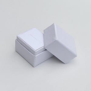 Puede incluir: Una caja de joyería blanca con tapa con bisagras, parcialmente abierta para revelar un interior blanco y suave. La caja es rectangular con un diseño limpio y minimalista. La tapa está separada, descansando junto a la caja abierta.