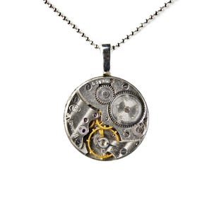 Puede incluir: Un collar con colgante de plata con un mecanismo de reloj circular. El colgante muestra intrincados engranajes, joyas y componentes met&aacute;licos. El collar tiene una cadena fina y una argolla plateada.