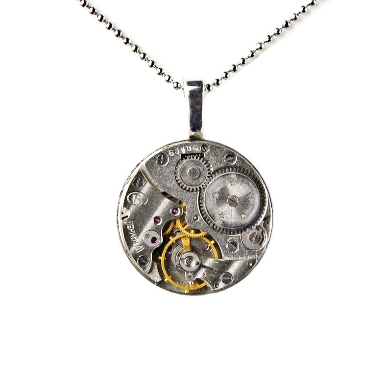 Puede incluir: Un collar con colgante de plata con un mecanismo de reloj circular. El colgante muestra intrincados engranajes, joyas y componentes met&aacute;licos. El collar tiene una cadena fina y una argolla plateada.