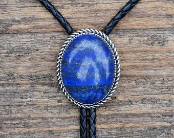 May include: A bolo tie with a blue lapis lazuli stone set in a silver oval pendant. The bolo tie has a black braided cord.