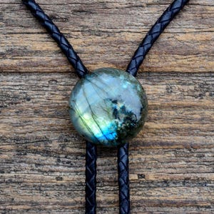 May include: A bolo tie with a round labradorite stone, displaying iridescent blue and green colours. The stone is set on a black braided leather cord. The background is a wooden surface, providing a rustic contrast.