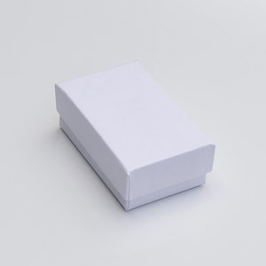 Puede incluir: Una peque&ntilde;a caja de regalo rectangular blanca con tapa. La caja est&aacute; hecha de un material liso y mate y est&aacute; colocada sobre un fondo blanco liso. La caja est&aacute; vac&iacute;a y lista para ser llenada.