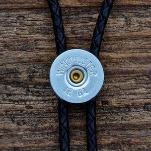 May include: A black braided leather bolo tie with a silver shotgun shell pendant. The shell is engraved with "WINCHESTER 12 GA".