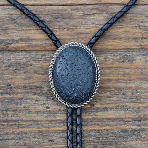 May include: A black bolo tie with a silver oval pendant featuring a dark gray stone. The stone has a rough, textured surface.