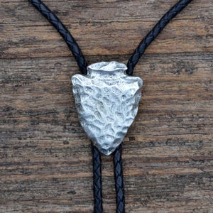 Puede incluir: Una corbata bolo plateada con un colgante de punta de flecha texturizada. El colgante está suspendido de un cordón de cuero trenzado negro. El fondo es una superficie de madera rústica.