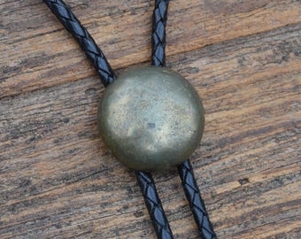 Puede incluir: Una corbata bolo con un gran colgante circular de color latón deslustrado. El colgante cuelga de dos cordones de cuero trenzado negro. La corbata descansa sobre una superficie de madera.