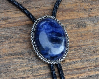 May include: A bolo tie featuring a large, oval, deep blue stone with silver-colored trim. The stone is set on a black braided leather cord. The tie is displayed on a wooden surface.
