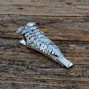 Könnte beinhalten: Eine silberfarbene Krawattenklammer mit einem detaillierten Farnblatt-Design. Die Klemme besteht aus Metall und hat eine strukturierte Oberfläche, wobei die komplizierten Details des Farns deutlich sichtbar sind. Die Klemme dient dazu, eine Krawatte zu halten.