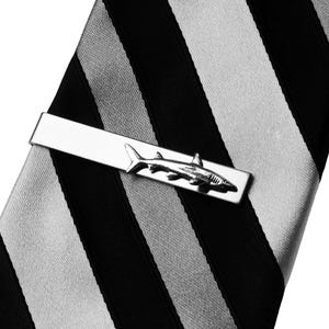 Puede incluir: Un pasador de corbata plateado con un diseño detallado de tiburón. El pasador se encuentra sobre una corbata a rayas negras, blancas y grises, creando un contraste llamativo. El tiburón está intrincadamente diseñado, agregando un toque de sofisticación.