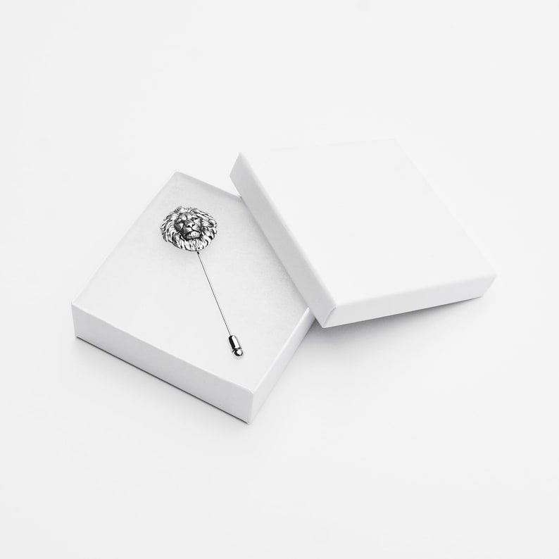 Puede incluir: Un broche de solapa de cabeza de le&oacute;n de plata en una caja blanca. El broche est&aacute; en un alfiler de plata y tiene un dise&ntilde;o detallado.