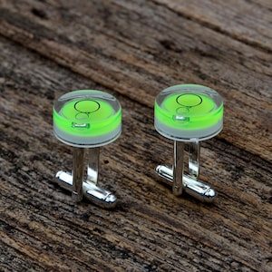 Può includere: Un paio di gemelli argentati dal design unico. Ogni gemello presenta una parte superiore trasparente e circolare con una bolla di livello verde brillante, montata su un perno d'argento con un'estremità arrotondata. Presentati su una superficie di legno rustico.