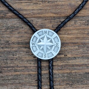 May include: A silver-colored bolo tie pendant with a compass rose design, featuring the letters N, E, S, and W. The pendant is attached to a black braided leather cord. The background is a wooden surface.