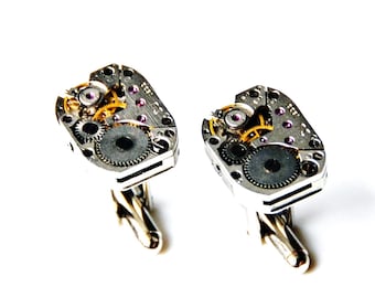 May include: A pair of silver cufflinks with intricate clockwork designs. The cufflinks feature exposed gears and small pink jewels.