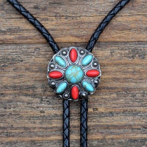 May include: A black leather bolo tie with a silver and turquoise bolo tie slide. The slide features a circular design with red and turquoise stones.