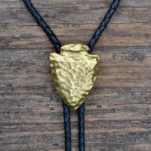 Puede incluir: Un bolo tie con una punta de flecha dorada y un cordón de cuero negro. La punta de flecha es texturizada y tiene un aspecto rústico.