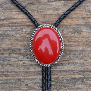 Könnte beinhalten: Ovale Bolo-Krawatte mit einem glänzenden roten Stein in der Mitte, umrahmt von einem silberfarbenen Seilmuster. Die Krawatte hat eine schwarze geflochtene Lederschnur. Der Hintergrund ist eine Holzoberfläche.