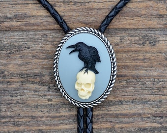 Puede incluir: Un lazo bolo con un cuervo negro posado sobre una calavera crema, sobre un fondo ovalado azul claro. El óvalo está enmarcado con un borde plateado. El lazo bolo tiene un cordón de cuero trenzado negro.