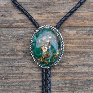 Peut inclure: Un bolo tie vert et or avec un pendentif ovale en argent. Le pendentif présente une pierre verte et or avec un motif tourbillonnant.