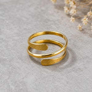 Op de afbeelding: Een goudkleurige metalen ring met een bypass-ontwerp met twee overlappende banden. De ring heeft een gladde, gepolijste afwerking aan de buitenranden en een licht getextureerd, geborsteld uiterlijk op de afgeronde uiteinden van de open ring.