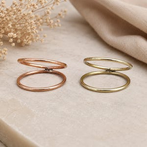 May include: Two thin metal rings, one in a copper tone and one in a gold tone. Each ring features a double-band design where two circular wires are joined at a single point, creating an open, stacked appearance with a smooth, polished finish.
